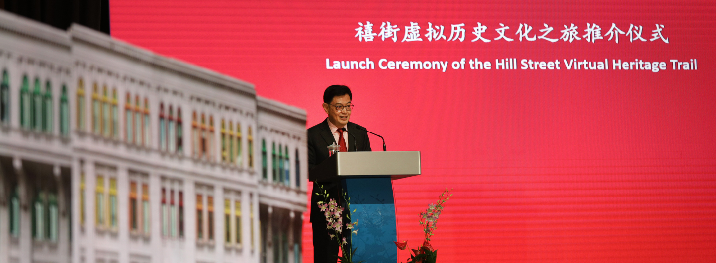 Man at podium with text: "Launch Ceremony of the Hill Street Virtual Heritage Trail." Blurred colorful building.